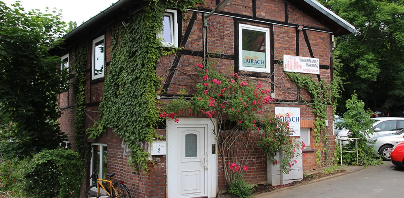 Das Bürogebäude in der Rudolf-Bultmann-Str. 2: ein berankter Backsteinbau mit Fachwerk und blühenden Rosen am Eingang